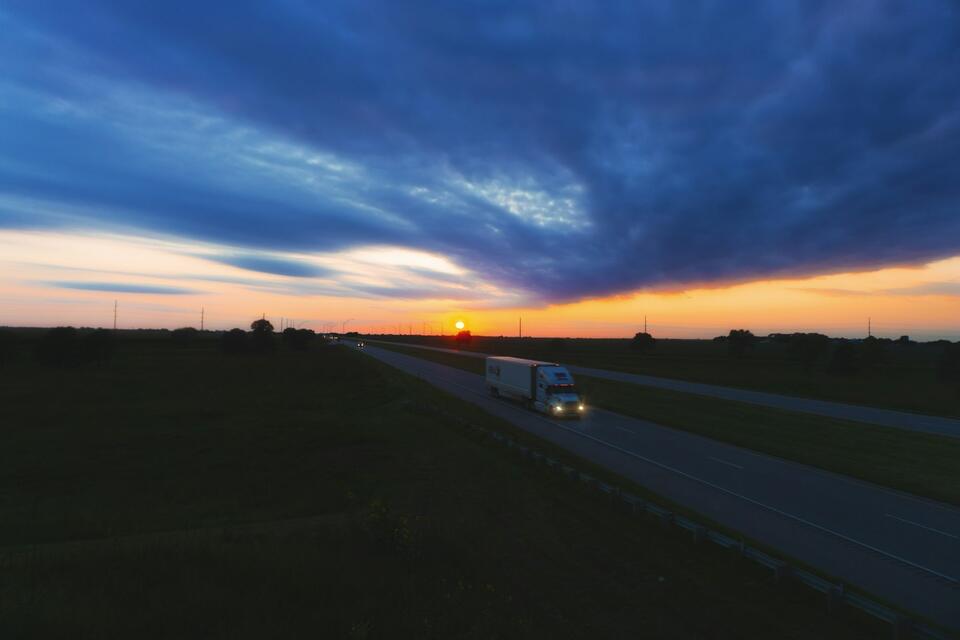 Semi truck with headlights on driving a long highway at sunset
